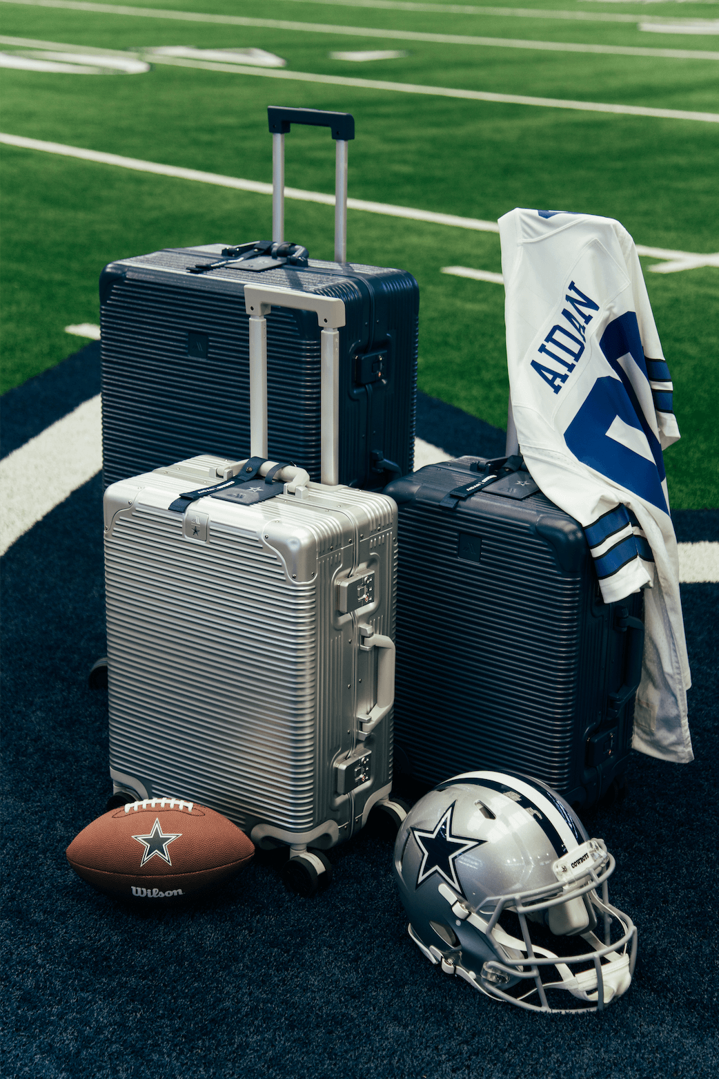 DALLAS COWBOYS X AIDAN collection featuring luggage, a jersey, a football, and a helmet on the field.