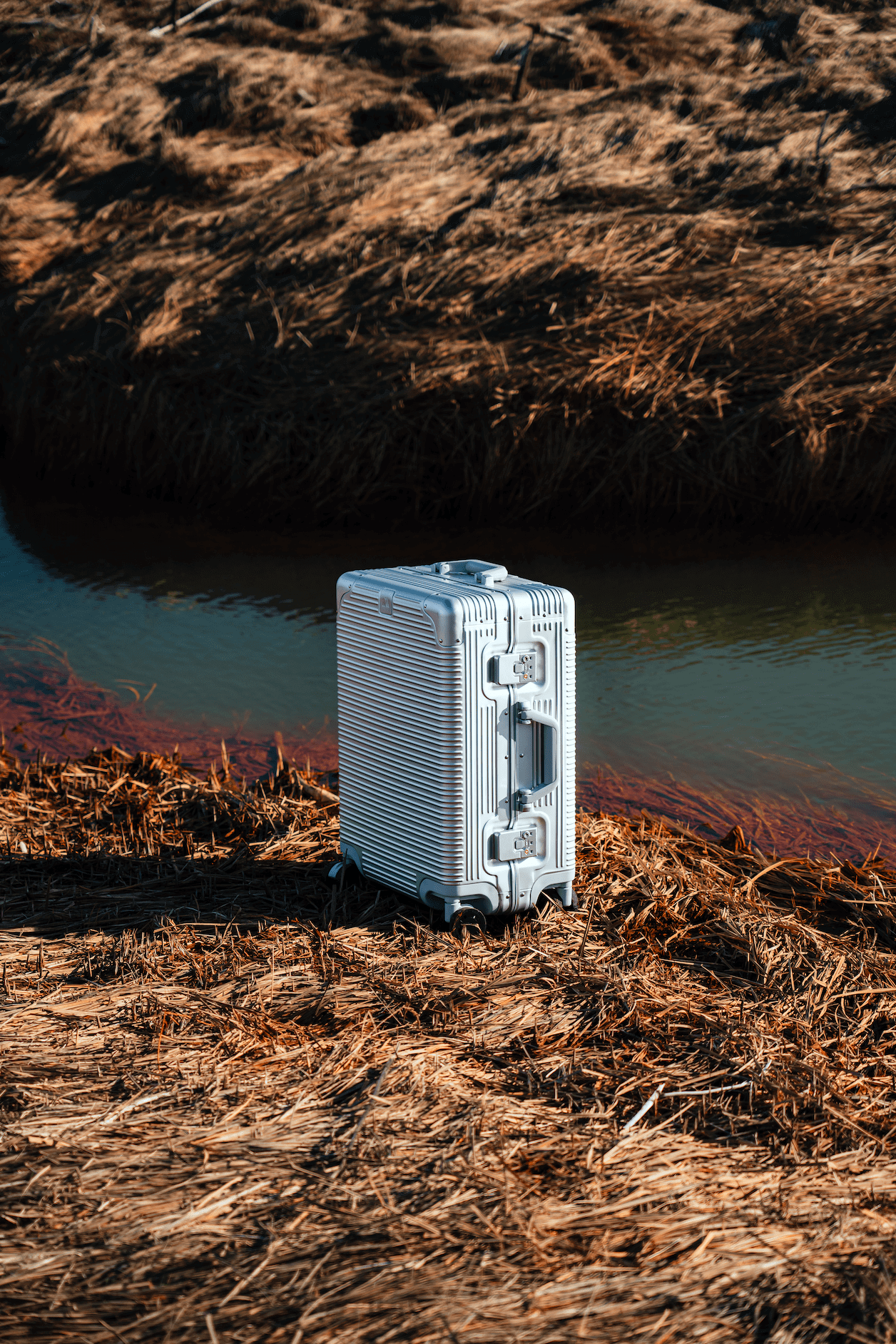 Stylish white suitcase on dry grass near a calm river, perfect for travel adventures.