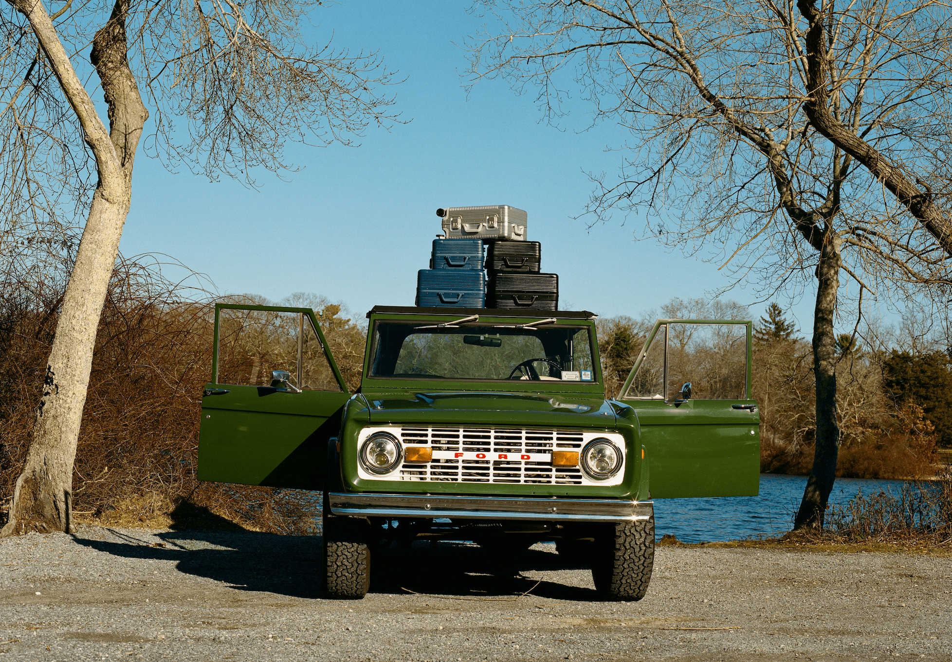 Classic green truck parked by the water with open doors and luggage on the roof, surrounded by trees.